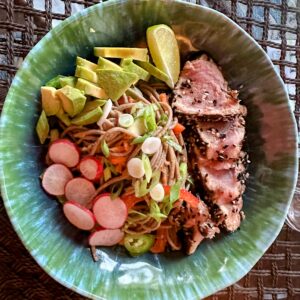 Soba noodle salad in a blue-green bowl topped with avocado, sliced radish, and seared tuna